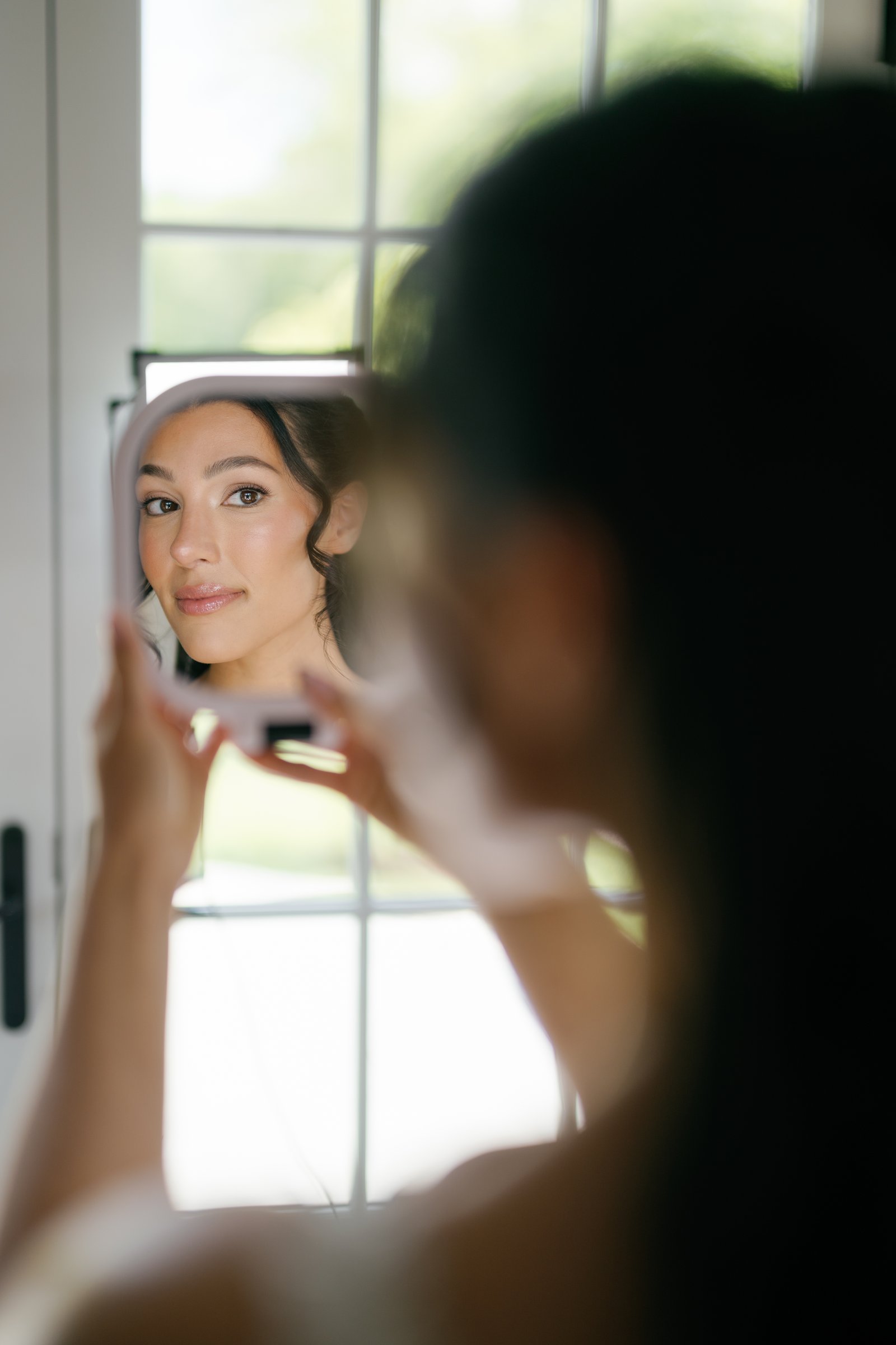 Kayla checking her makeup in a hand mirror by the window