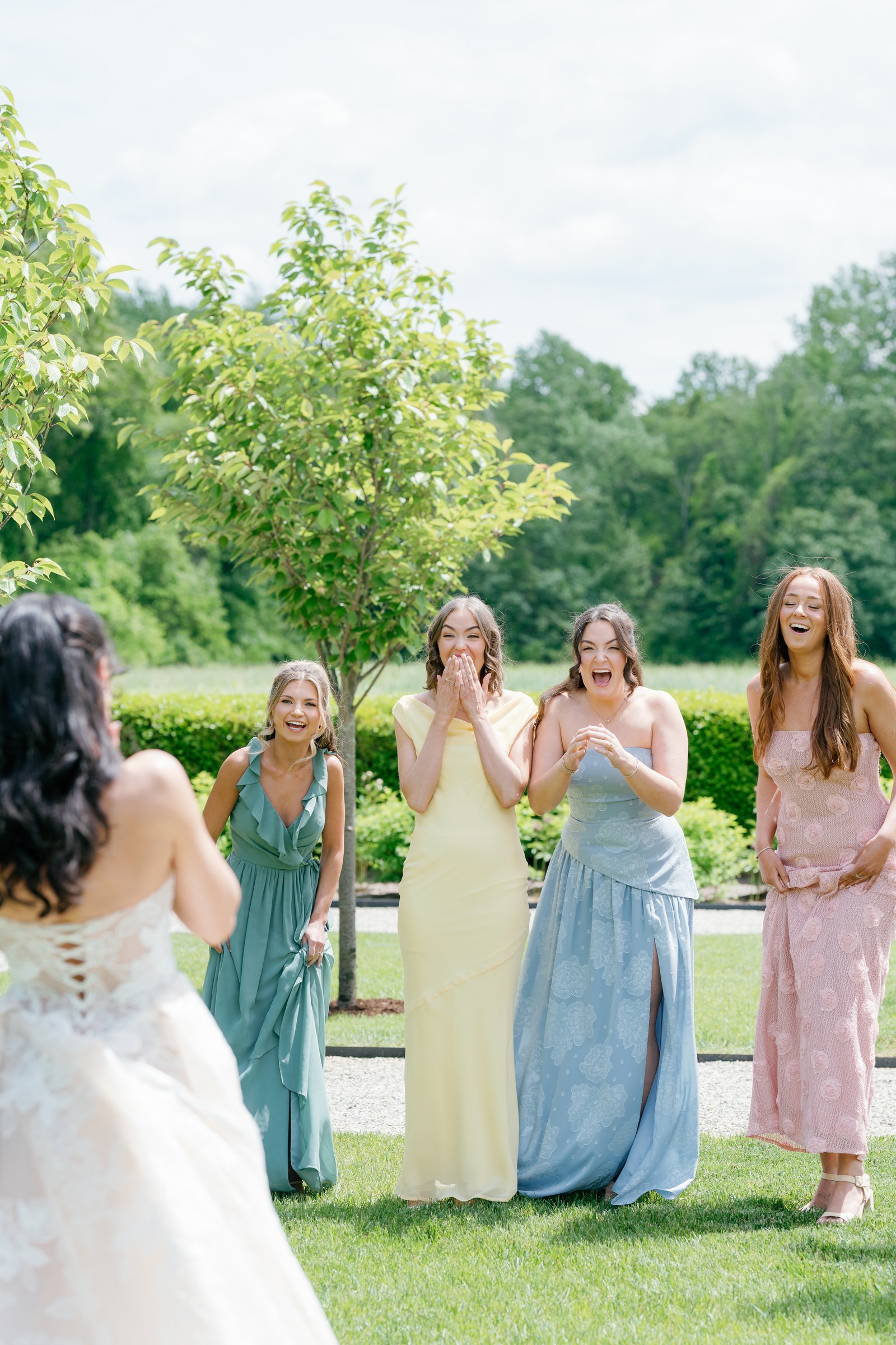Kayla's bridesmaids in pastel gowns reacting as they see her in her dress on the lawn
