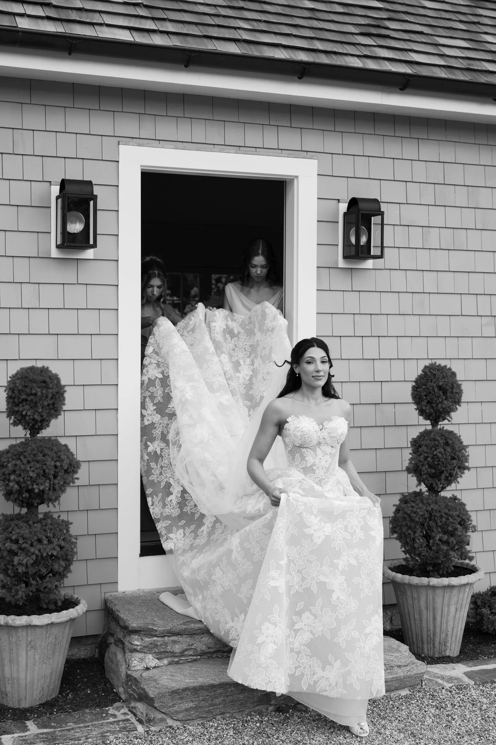 Kayla stepping out of the cottage door between two topiaries as a bridesmaid lifts her train, in black and white