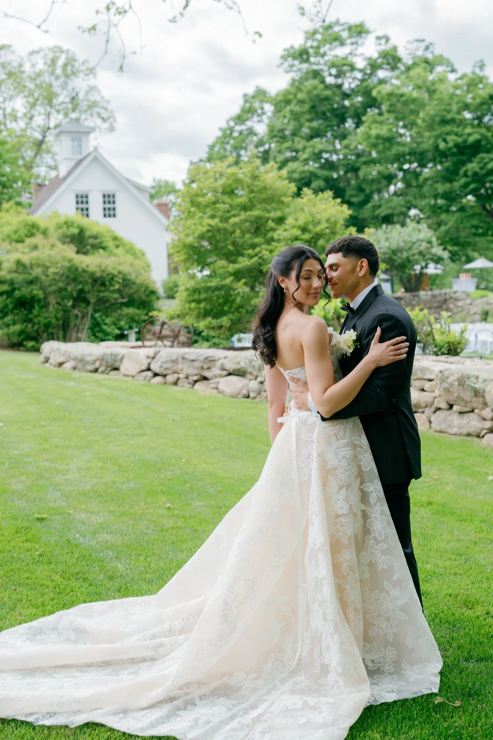 Kayla and Avery holding each other on the lawn by the stone wall, her train behind her