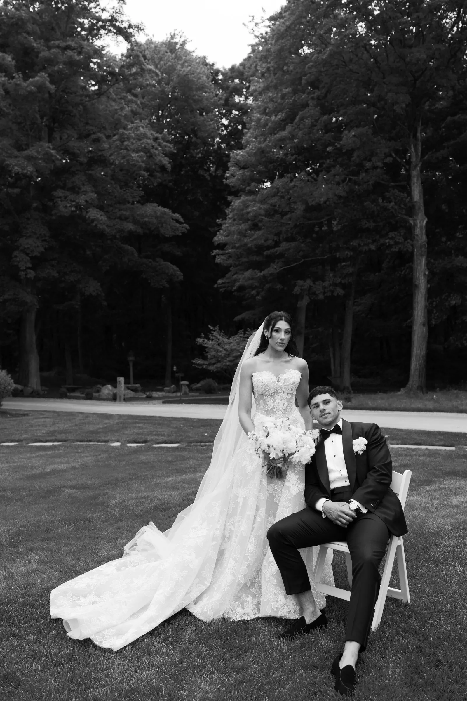 Kayla standing beside Avery as he sits on a chair on the lawn, in black and white