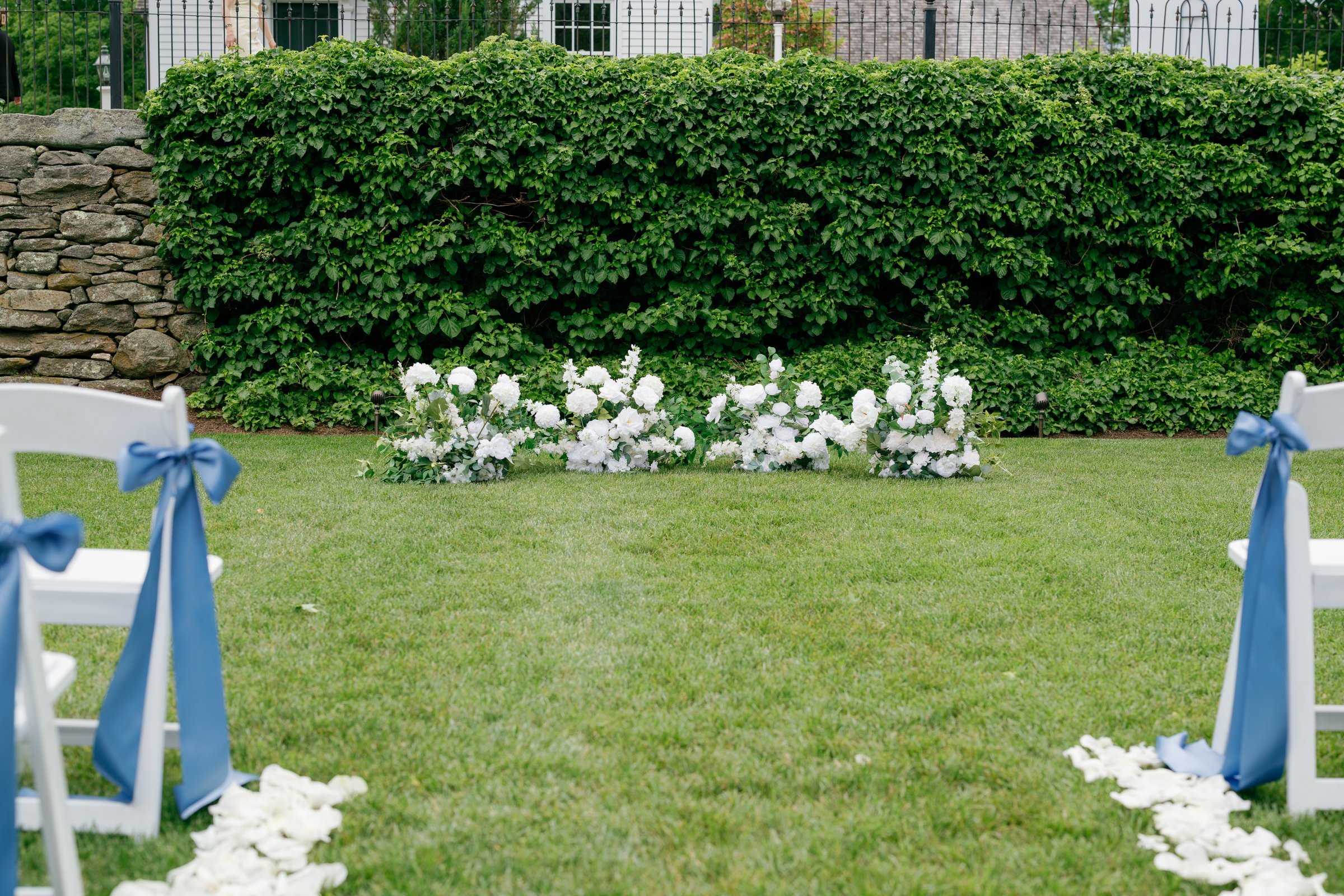 The ceremony arch of white flowers on the lawn, with blue ribbons on the aisle chairs